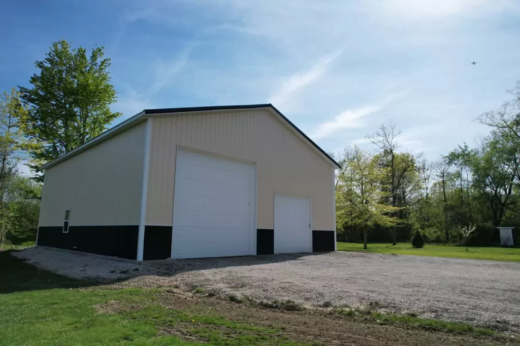 Wide front view of post-frame garage with white overhead doors and black wainscot, surrounded by trees in Amherst NY.