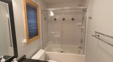 Before image of a bathroom ready for upgrade, featuring a traditional bathtub with a shower and clear glass sliding door, light beige tiles with diamond accents, wooden-framed window with blue blinds, and white walls, exemplifying a typical Stately Kitchen and Bath renovation project.