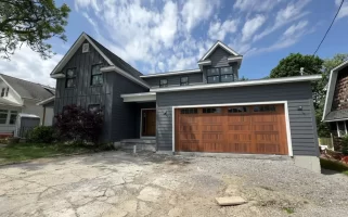 Buffalo home addition with modern gray siding, wood garage door, and second-story expansion under a bright summer sky.
