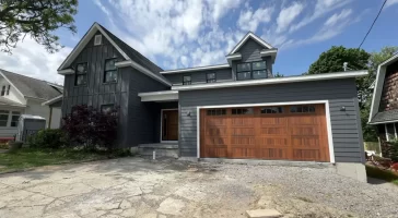 Buffalo home addition with modern gray siding, wood garage door, and second-story expansion under a bright summer sky.