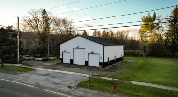 Durable white and black post-frame structure by Stately Pole Barns in Holland, NY, featuring three garage doors and a gravel driveway.
