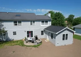Exterior view of a completed home addition, featuring a new in-law suite with gray siding and a black roof, connected to the main house. The backyard includes a patio area with outdoor furniture and a grill, set against a clear blue sky.