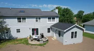 Exterior view of a completed home addition, featuring a new in-law suite with gray siding and a black roof, connected to the main house. The backyard includes a patio area with outdoor furniture and a grill, set against a clear blue sky.