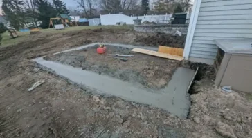 Foundation being laid for a home addition, showing early construction stages with fresh concrete, adjacent to an existing house structure, in a residential backyard setting.