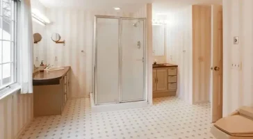 Before photo of an outdated bathroom showing a shower stall, double vanity, striped wallpaper, and hexagonal tile flooring with beige tones.