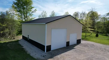 Corner view of custom post-frame garage with black metal roof and dual overhead doors in wooded Amherst NY setting.