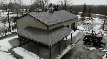 Elevated view of a spacious pole barn with open garage doors and a covered lean-to, with a snowy landscape and construction equipment in the foreground.